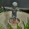 Auf einem Brunnen aus Kalkstein steht eine Figur. Die Figur zeigt einen kleinen nackten Jungen. Er hält zwei Fische in der Hand. Aus den Mäulern der Fische kommt jeweils ein Wasserstrahl.