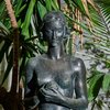 Die Bronzefigur einer Frau steht vor Palmen. Sie ist nicht bekleidet und hält einen Apfel in der Hand. Sie stellt die biblische Figur Eva dar.
