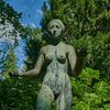 Eine Bronzefigur kniet mit ihrem linken Bein auf einem kleinen Podest. Die Figur stellt eine Frau dar. Sie trägt keine Kleidung.
