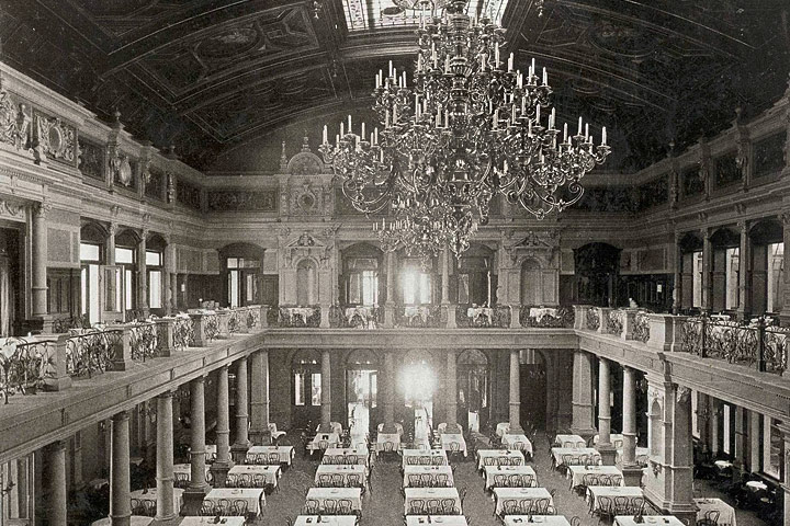 Eine geschichtliche Abbildung des Gesellschaftshauses, das sich am Palmenhaus anschließt. In der Halle stehen kleine Tische mit Stühlen und von der Decke hängt ein prunkvoller Kronleuchter. 